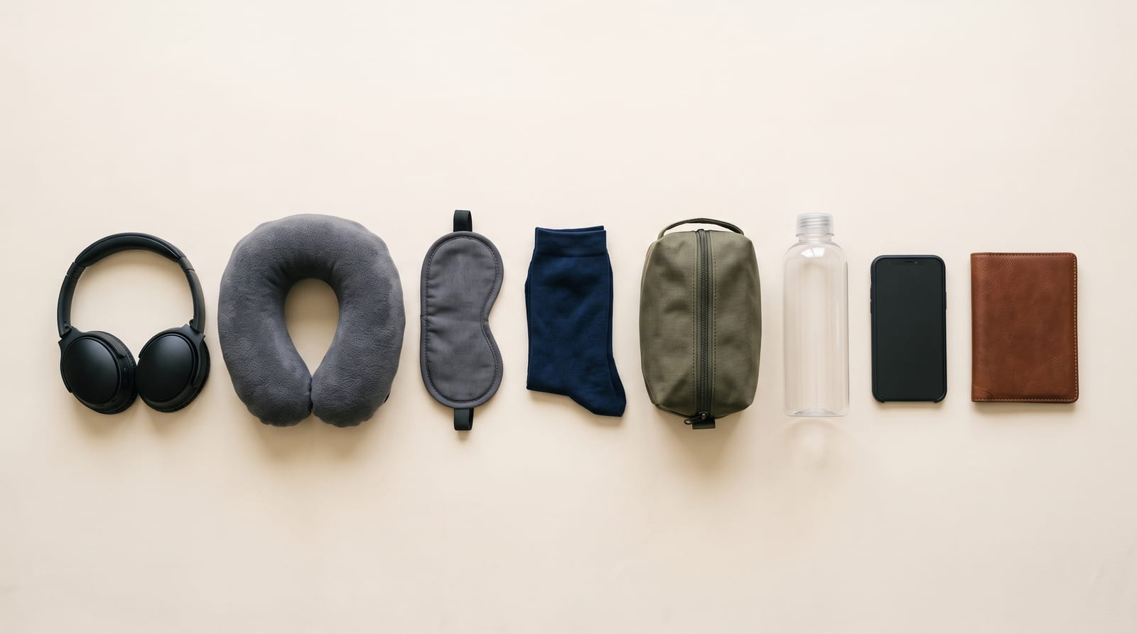 Flat-lay of airplane travel essentials on a cream background — noise-canceling headphones, gray neck pillow, sleep mask, navy socks, toiletry bag, water bottle, phone, and a leather passport wallet