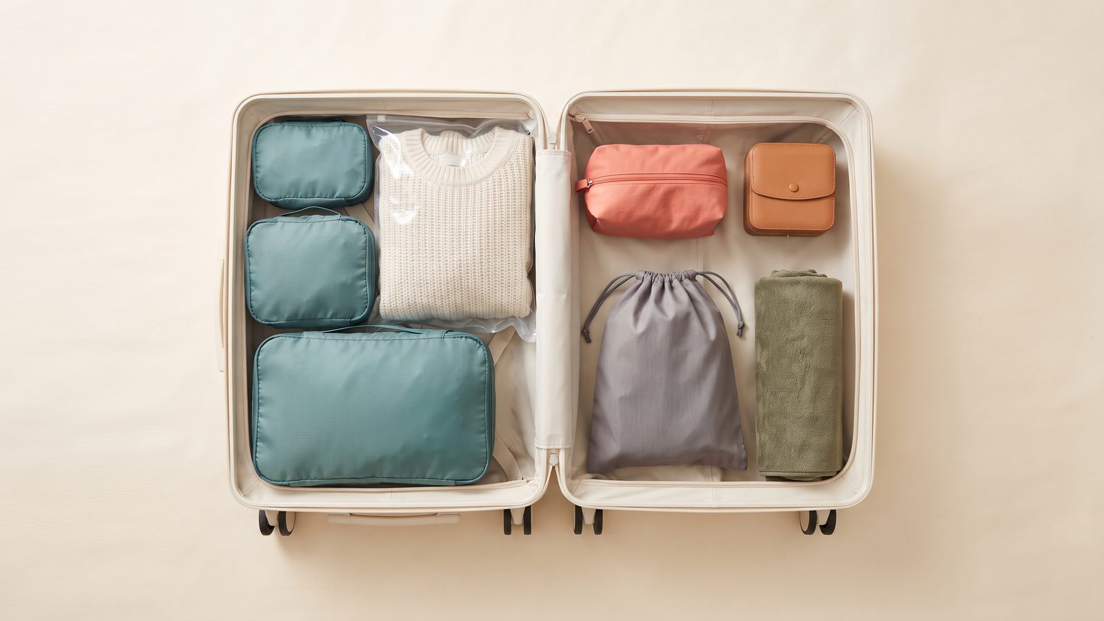 Top-down view of an open cream hardside suitcase packed neatly with teal packing cubes in three sizes, a clear compression bag with a folded sweater, a coral toiletry pouch, a tan jewelry case, a gray shoe bag, and a rolled olive towel on a cream background