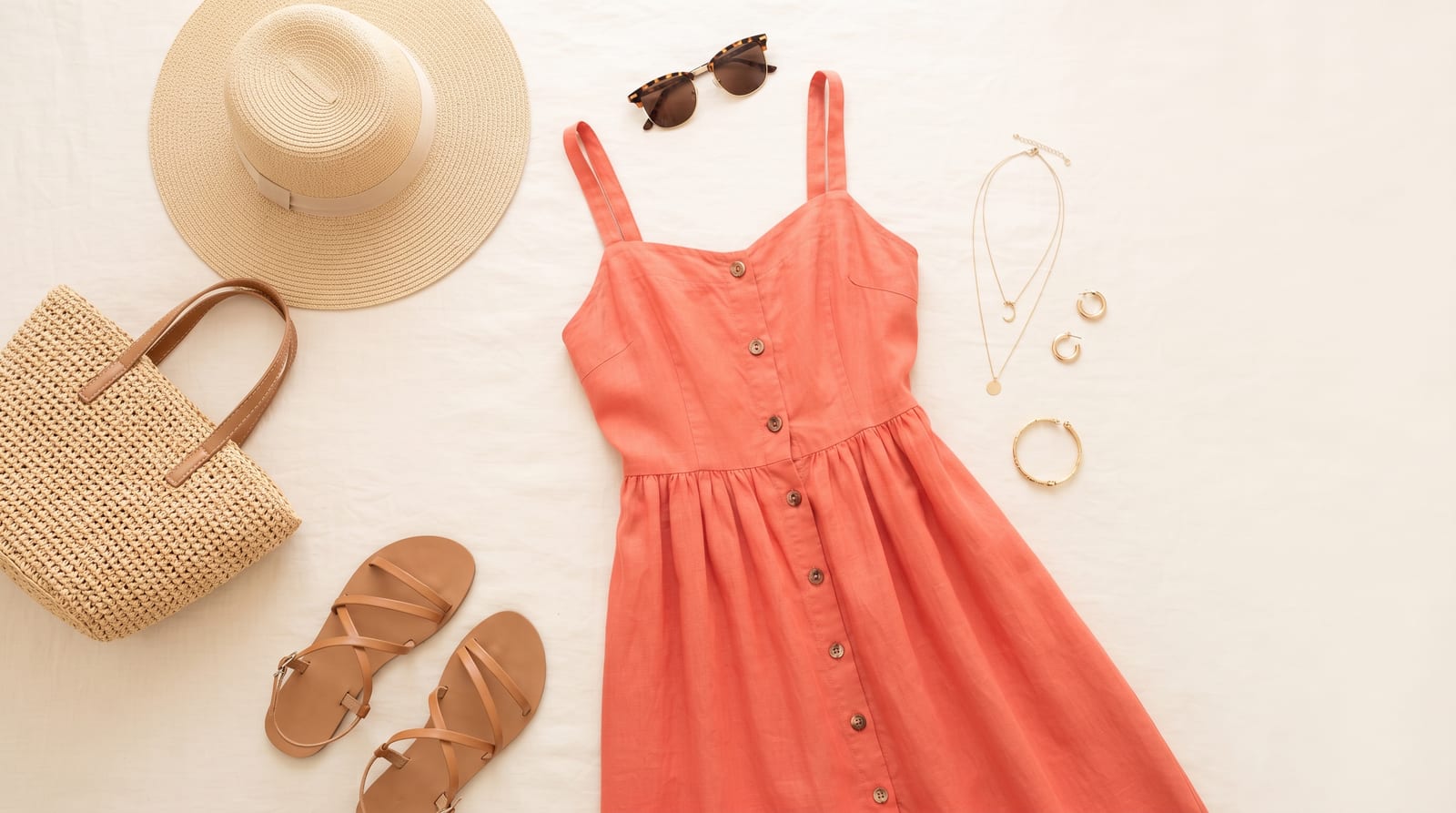 Vacation outfit flat-lay — coral button-front sundress, wide-brim straw hat, woven tote, strappy tan sandals, sunglasses, and delicate gold jewelry on a cream background